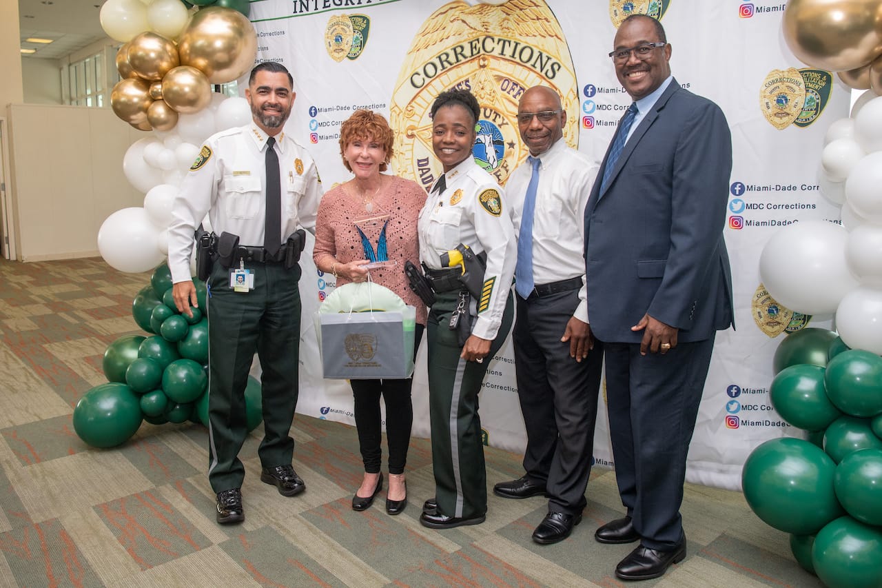 Photo of (from left to right) Director James Reyes, Lucy Collins, Deputy Director Cassandra Jones, Counselor Terrence Matthews, and Chief of Corrections JD Patterson, the staff of Miami-Dade Correctional Center, happily standing in front of a photo wall. Lucy holds the volunteer award she received from the Center.