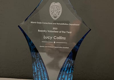 Photo of the beautiful Reentry Volunteer of the Year Award 2023. It is diamond-shaped, etched glass with blue translucent sides and is awarded to Lucy Collins by Miami-Dade Corrections and Rehabilitation Department.