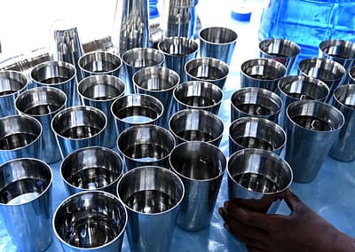 Dozens of shiny, metal cups filled with clean drinking water for schoolchildren in Bantoli, India, sit on a table covered with a clear plastic. They water and cups were delivered by Food for People staff.