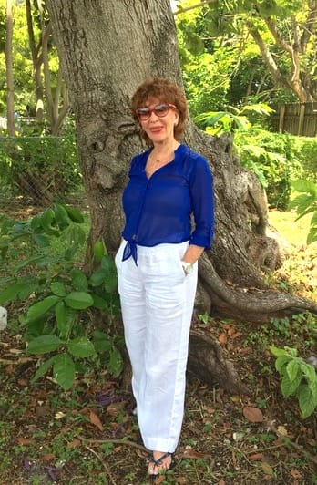 Photo of Lucy Collins, Peace Education facilitator and Reentry Volunteer of the Year Awardee, poses in front of a large tree in a lush, green Florida park. She is wearing sunglasses, a long-sleeved royal blue blouse with long white linen pants.