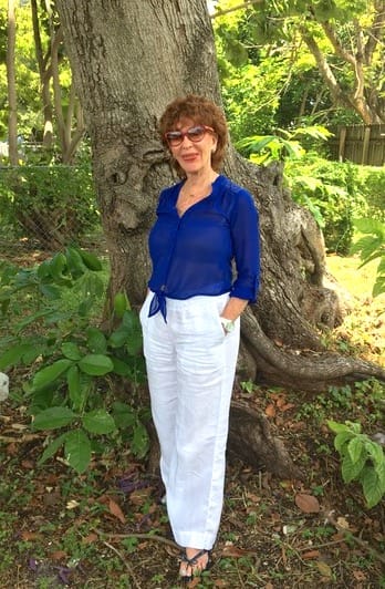 Foto de Lucy Collins, facilitadora de Educación para la Paz y ganadora del Premio al Voluntario de Reingreso del Año, posa frente a un gran árbol en un exuberante y verde parque de Florida. Lleva gafas de sol, una blusa azul real de manga larga con pantalones largos de lino blanco.