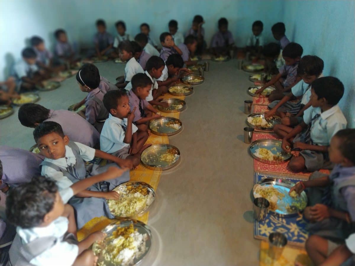 Jóvenes escolares de Bantoli, India, se sientan sobre coloridas esteras en una sala para disfrutar de un abundante y delicioso almuerzo de arroz, daal y verduras entregado y servido por el equipo de Food for People.