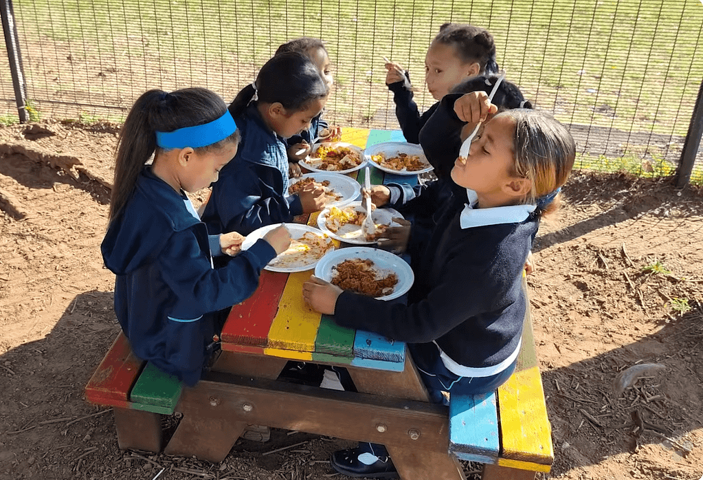 Six young schoolgirls sit on a rainbow colored wooden table in the school yard and enjoy the meals provided by the newly renovated Food for People kitchen in Cape Flats, South Africa.