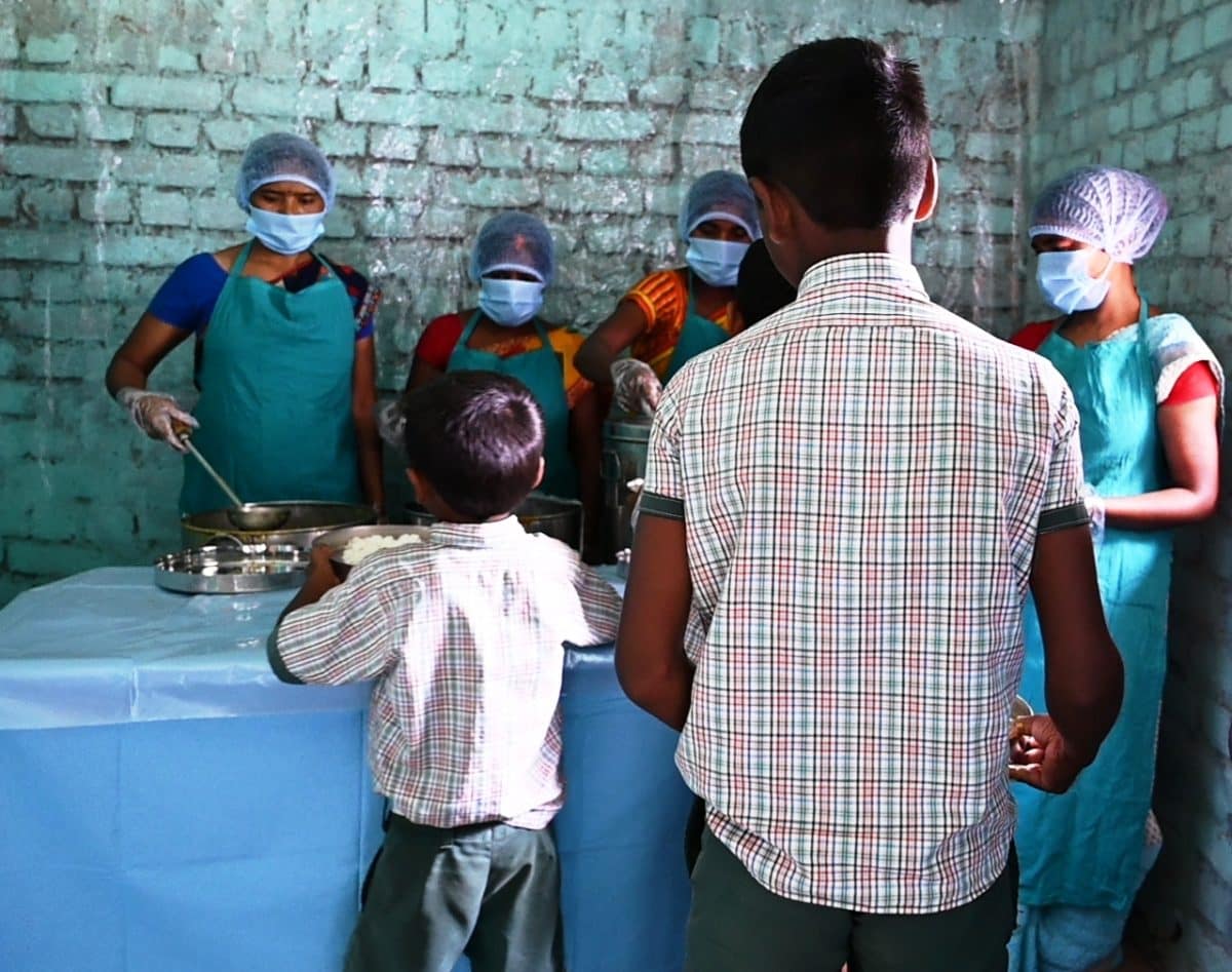 Dos jóvenes escolares llevan sus bandejas de comida a los camareros, quienes usan mascarillas, protectores para cabeza y ropa además de guantes, lo que ayuda a mantener altos estándares sanitarios.