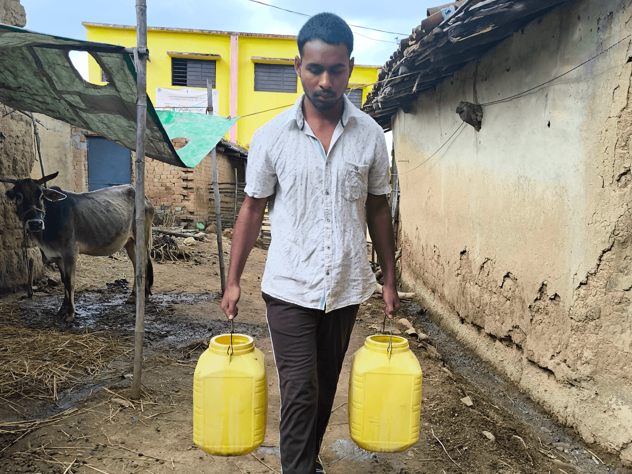 Yashwant trasporta due contenitori d'acqua gialli mentre passa accanto alla sua mucca e ad alcune case di mattoni di fango.